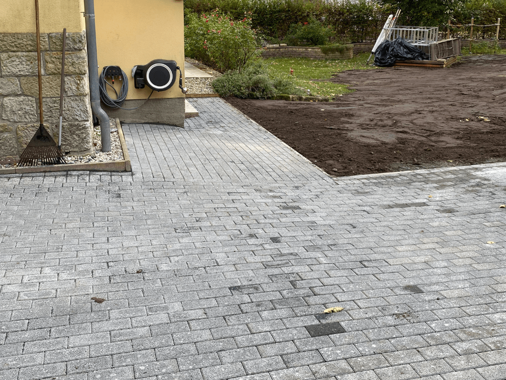 Neu gepflasterter Hofbereich mit grauen Steinen, angrenzend an ein gelbes Haus und eine vorbereitete Gartenfläche.