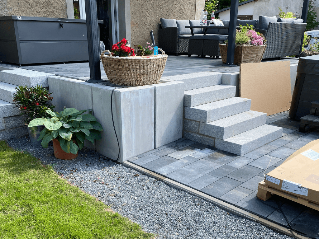 Terrasse mit grauen Steinfliesen, Treppe aus Betonstufen und dekorativen Pflanzen, angrenzend an eine Sitzgruppe mit Gartenmöbeln.