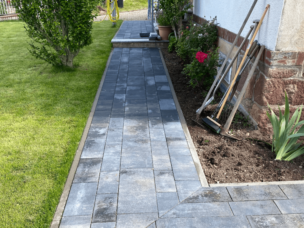 Gartenweg aus grauen Pflastersteinen, der an einem gepflegten Rasen und einem Blumenbeet entlangführt, mit Gartenwerkzeugen an der Hauswand.