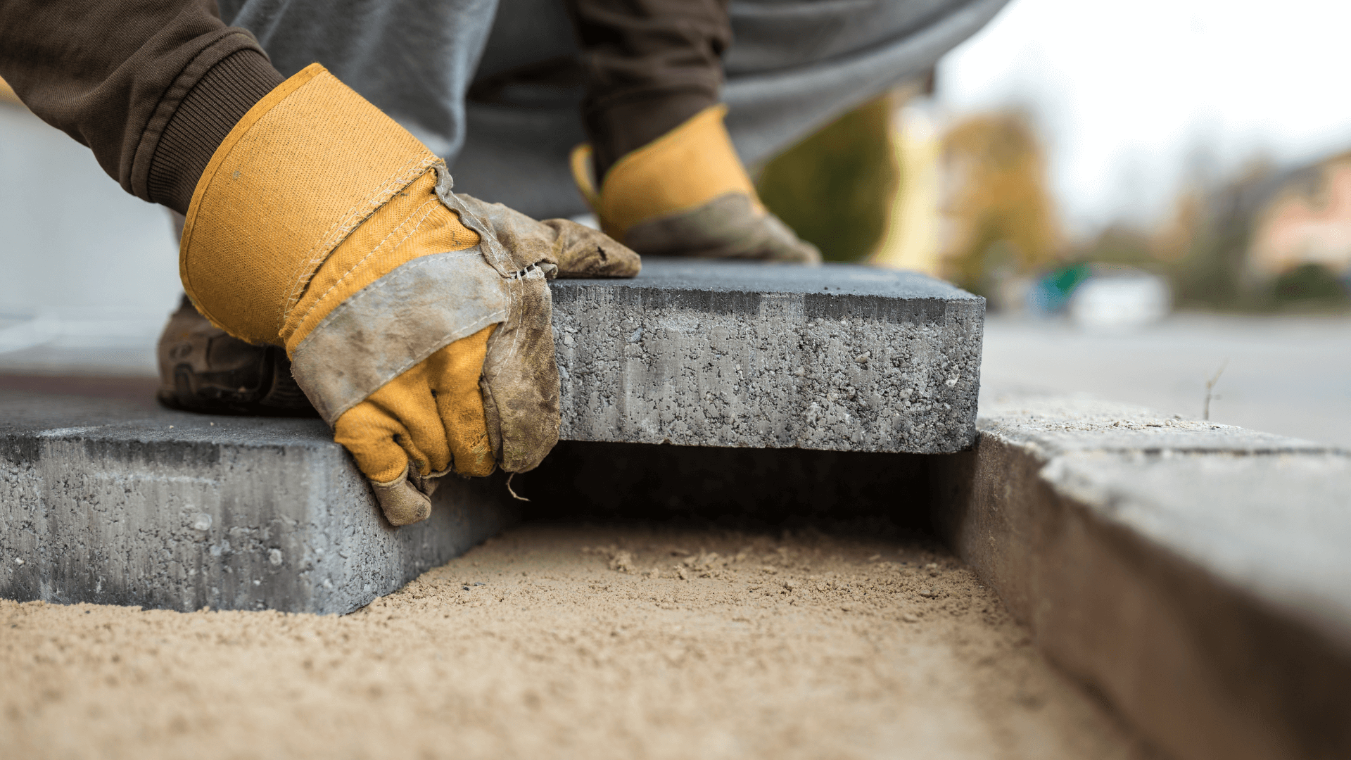 Pflasterarbeiten mit grauen Steinen, ein Arbeiter setzt mit Handschuhen einen Stein in ein Muster ein, während ein Hammer am Boden liegt.