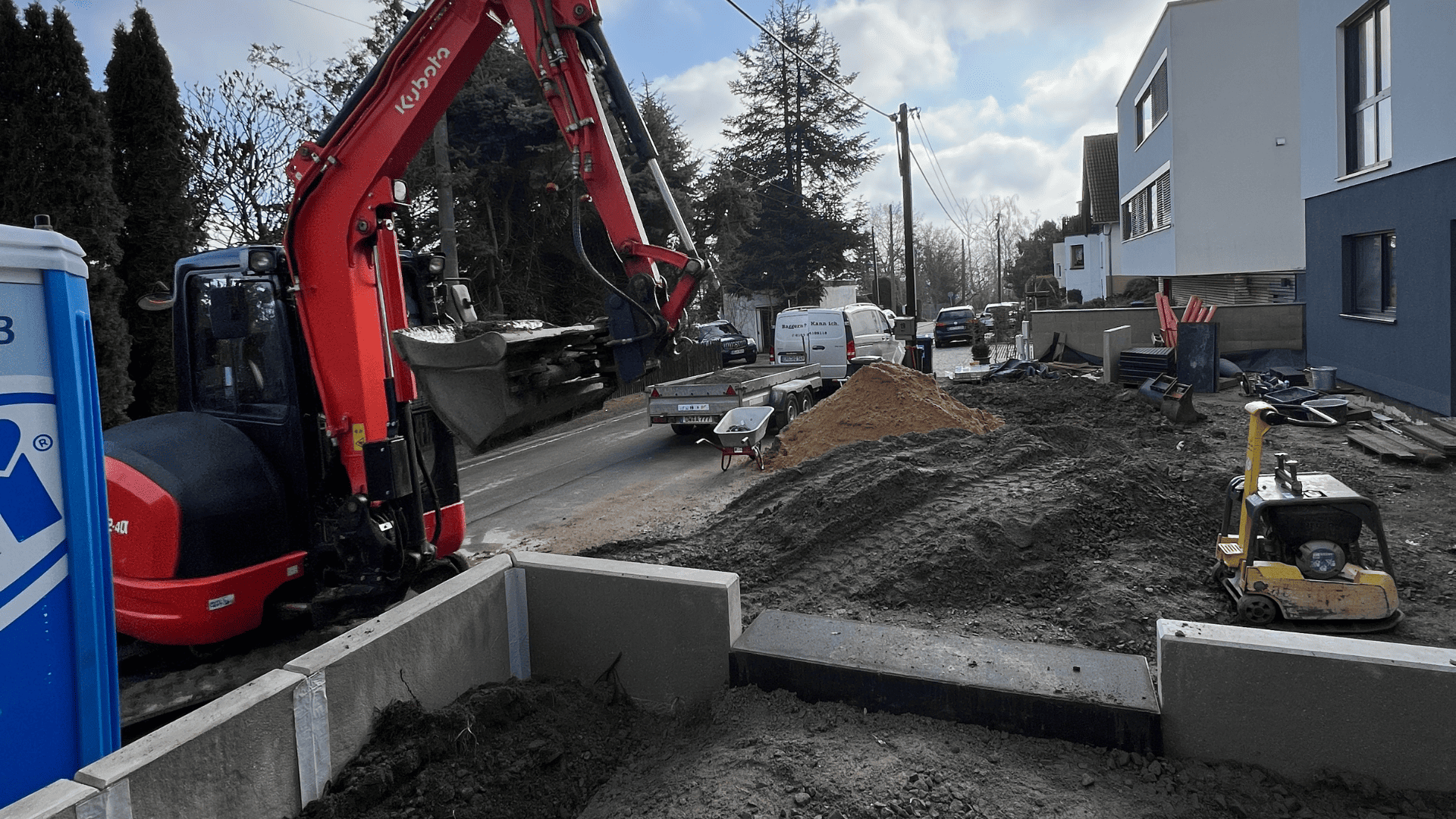 Baustelle im Bereich Galabau mit Bagger, Schubkarre und Baumaterialien vor einem modernen Wohnhaus.
