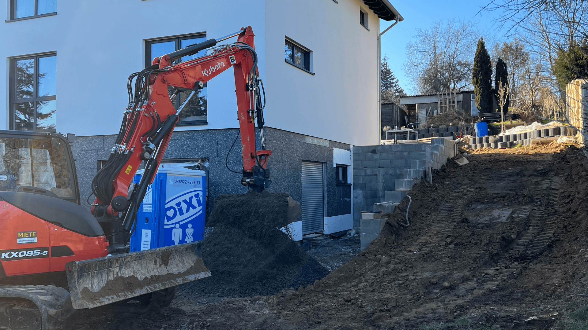 Ein Minibagger verteilt Schotter vor einem modernen Einfamilienhaus, während im Hintergrund eine neue Hangbefestigung mit Betonsteinen errichtet wird.